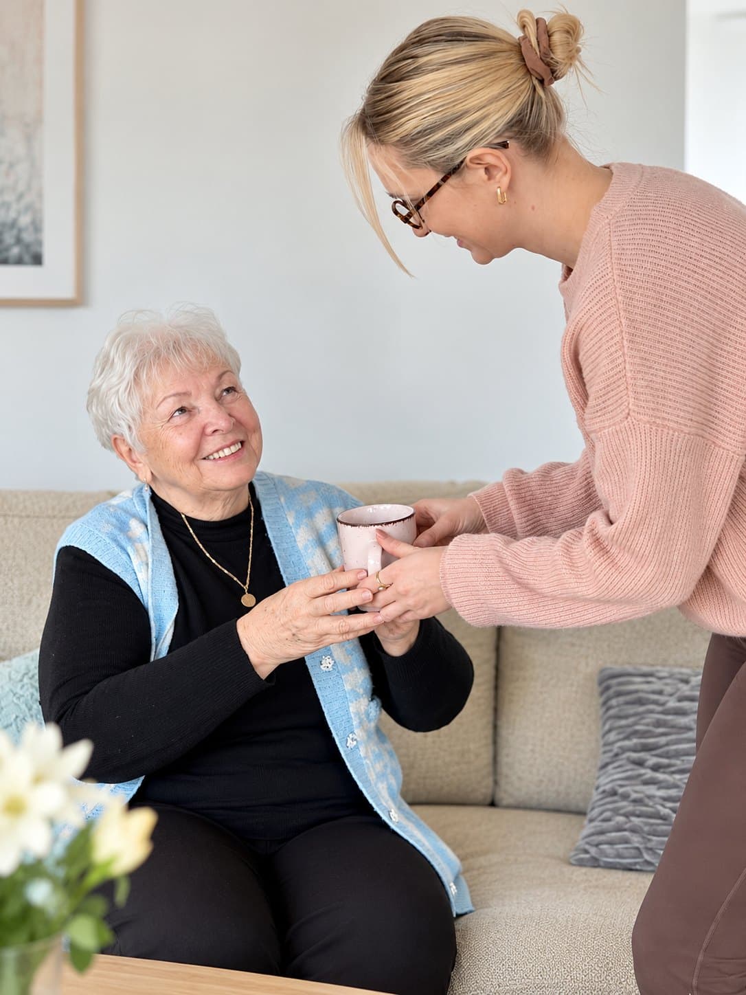 Betreuungskraft reicht einer Seniorin auf dem Sofa eine Tasse Tee